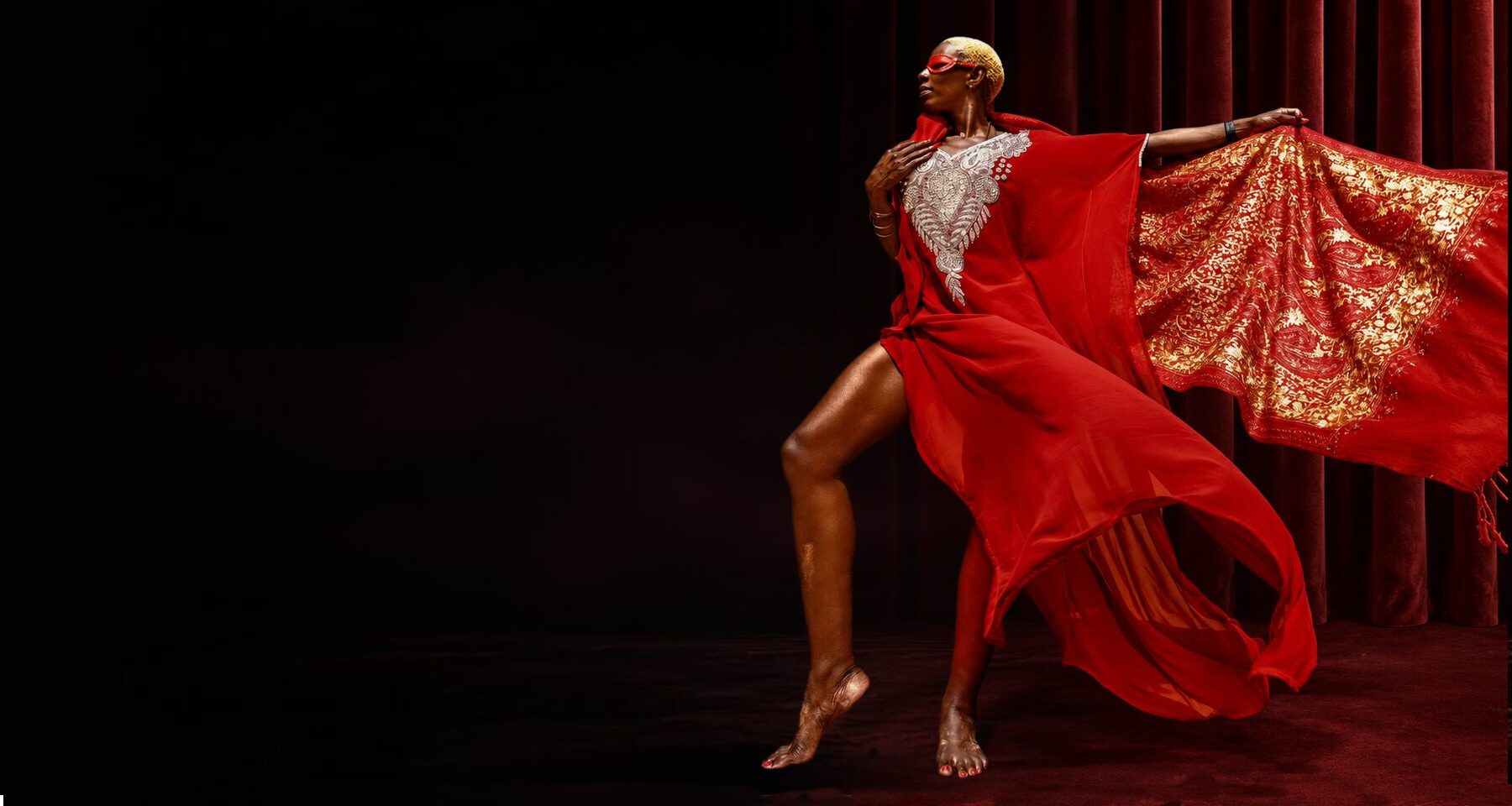 The Fit Goddess wearing a red mask and red and gold gown before burgundy curtains
